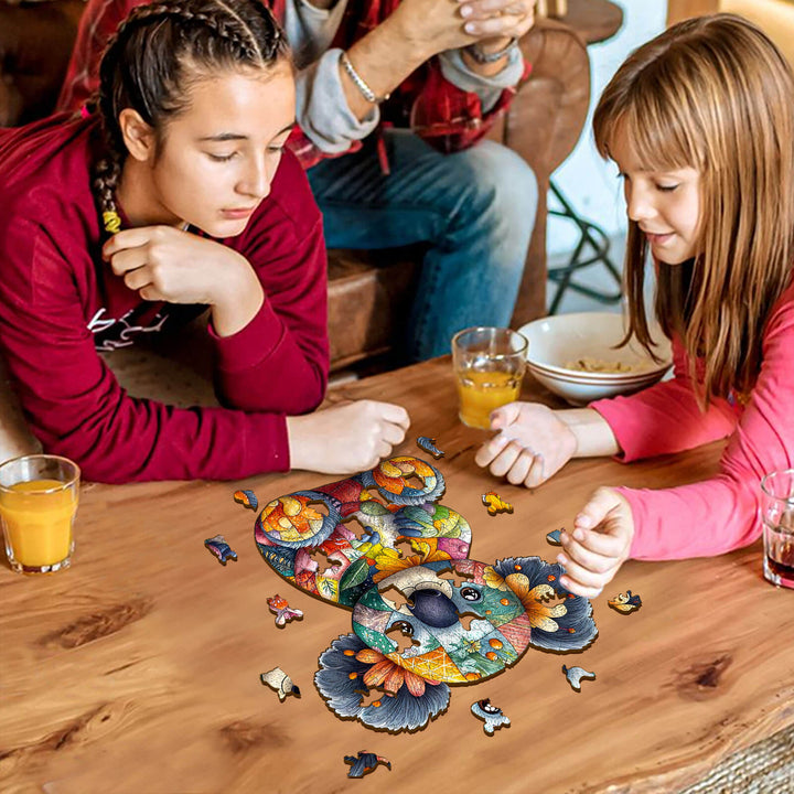 Two children engaged in solving a colorful Patchwork Koala wooden jigsaw puzzle on a wooden table.
